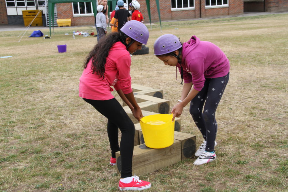 Two young girls wearing helmets work together to carry yellow bucket on team building activity
